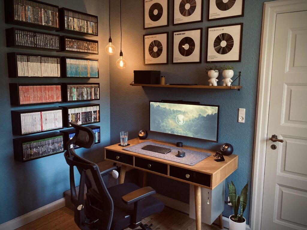 Minimalist gaming and workstation setup with a curved ultrawide monitor, warm hanging lights, wall-mounted video game collection shelves, and framed vinyl-style artwork.