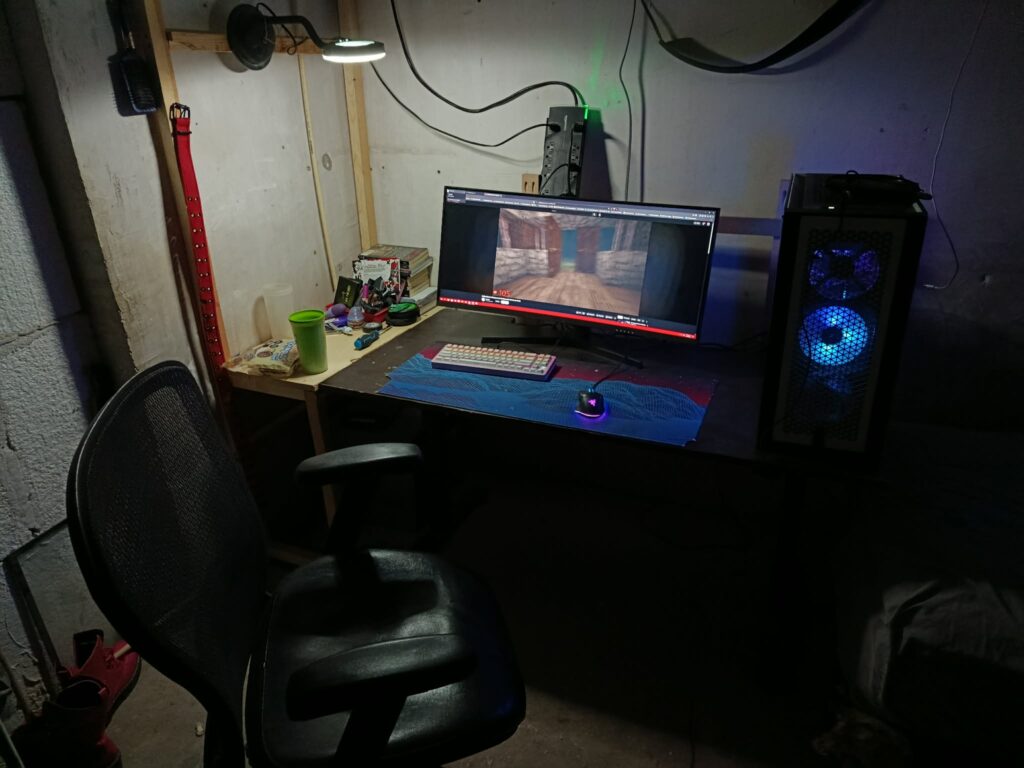 Dark basement gaming setup with ultrawide monitor, RGB gaming PC, and exposed unfinished walls.