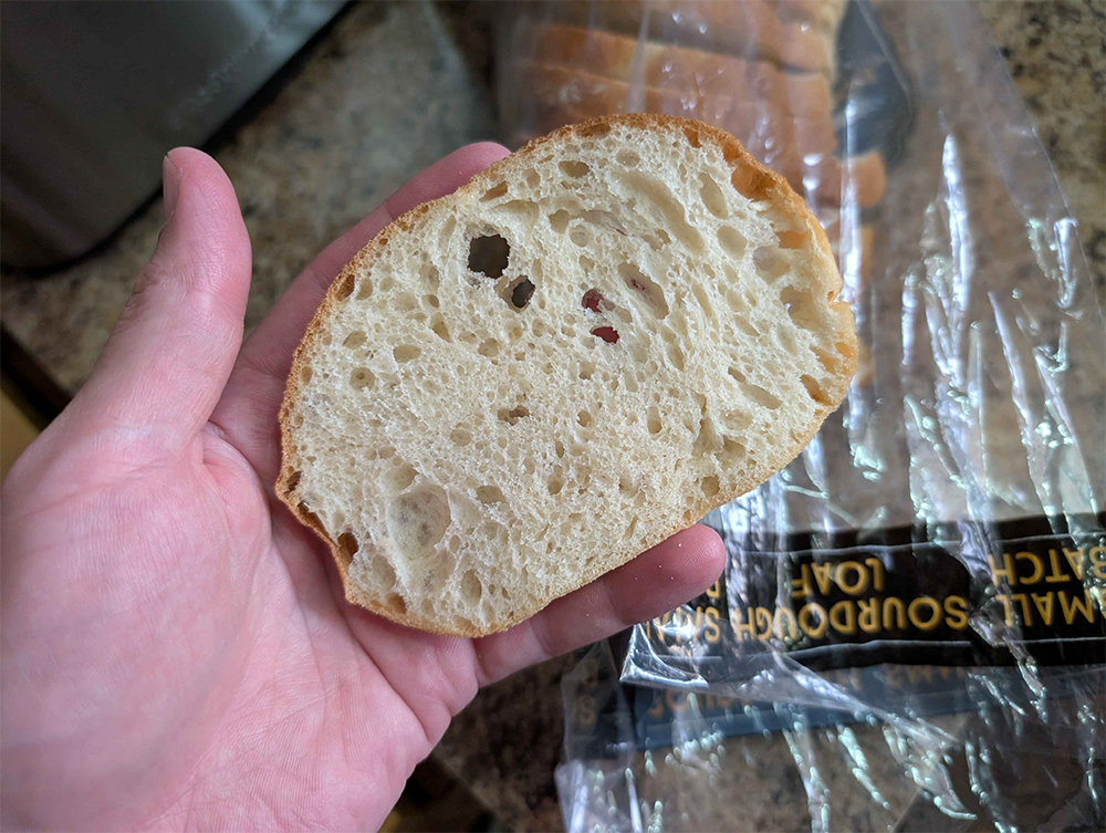 Small slice of Aldi Small Batch Sourdough Bread held in a person’s hand, with the loaf packaging visible in the background.