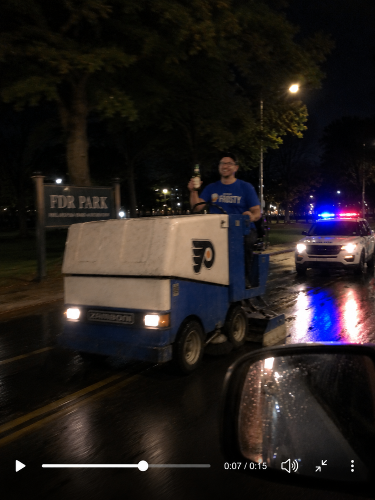 Blurry cellphone-style nighttime image showing Frosty Mugg driving a Zamboni near FDR Park while holding a beer as police vehicles follow behind him.
