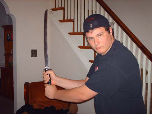 A young man wearing a backwards baseball cap holds a large machete-style knife while posing inside a house near a staircase.