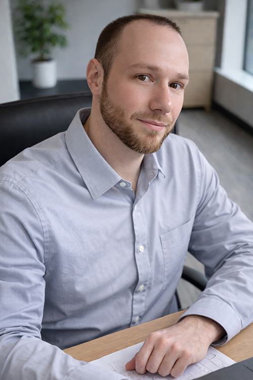 A man with a short beard and closely shaved hair sits at a desk wearing a light blue button-up shirt while looking toward the camera.